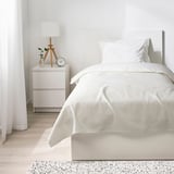 Modern hálószoba fehér STENFRÖMAL ágytakaróval, egyszemélyes ágyon, világos fa éjjeliszekrényrel, lámpával, vázával és órával. Tágas és minimalista kialakítás.