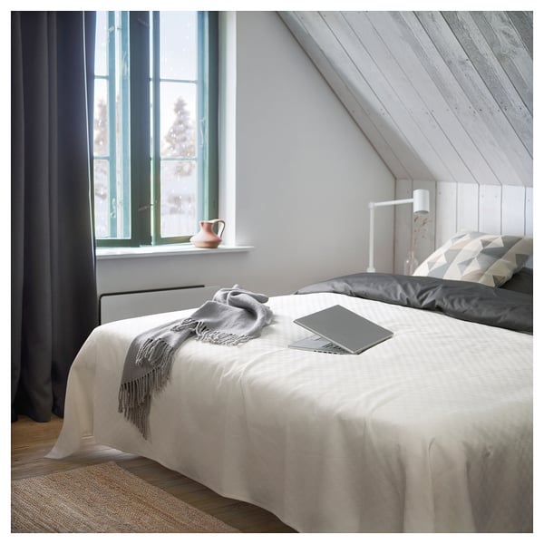 Modern hálószoba fehér ágytakaróval, szürke takaróval, párnákkal, laptoppal, lámpával és nagy ablakkal, amelyen fák láthatók.