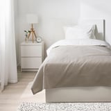 Bézs ágytakaró, párnák, éjjeliszekrény lámpával, váza, óra. Minimalista, hangulatos szoba.