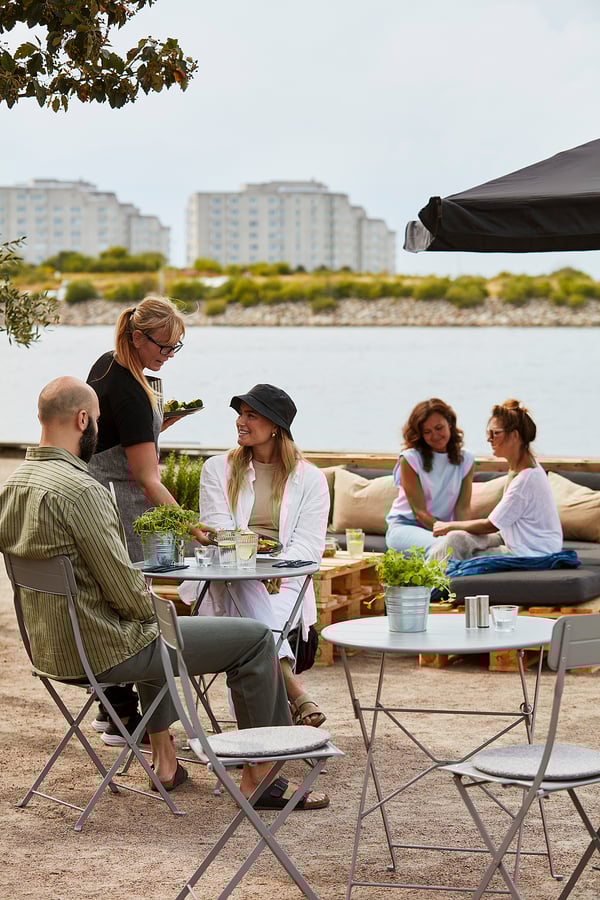 Öt ember vacsorázik a szabadban, a vízen, egy esernyő alatt, a háttérben épületekkel és zölddel.