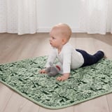 Baba erdei témájú szőnyegen, hason fekve, felfedezve a plüssjátékot.