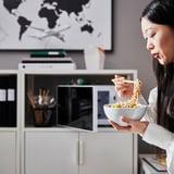 Nő tésztát eszik a TROTTEN fehér tárolóasztalánál. Az íróasztal állítható lábakkal, zárható tárolóval és fémcső szerkezettel rendelkezik.