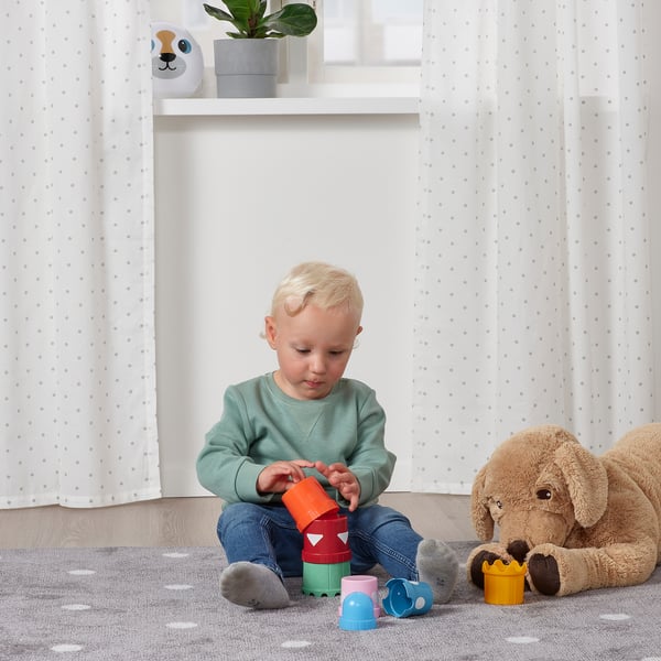 Kisgyermek színes egymásra rakható csészékkel játszik a padlón, puha játékokkal körülvéve.