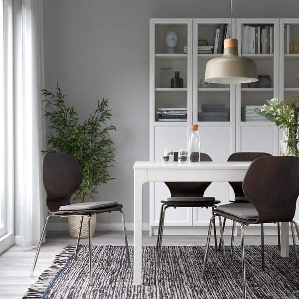 Minimalist dining area with white table, dark chairs, large window, potted plant, bookshelf, and pendant light.