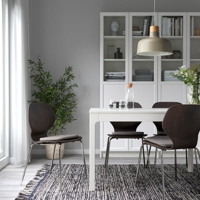 Minimalist dining area with white table, dark chairs, large window, potted plant, bookshelf, and pendant light.