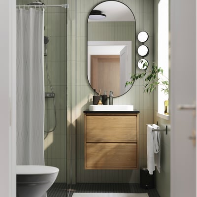 Modern bathroom with light green tiles, oak cabinet, white sink, round mirror, glass shower, and towel rack.