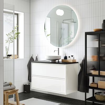 Modern bathroom with ÄNGSJÖN white washstand, round basin, wall-mounted, sleek design, multiple drawers, large mirror, black & white tiles.
