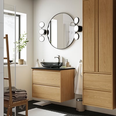 Modern bathroom: oak cabinet, black sink, round mirror, lights. Towels right.
