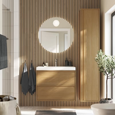 Modern bathroom featuring ÄNGSJÖN oak vanity, round mirror, & plants. Vanity stands tall, drawing focus amidst sleek decor.