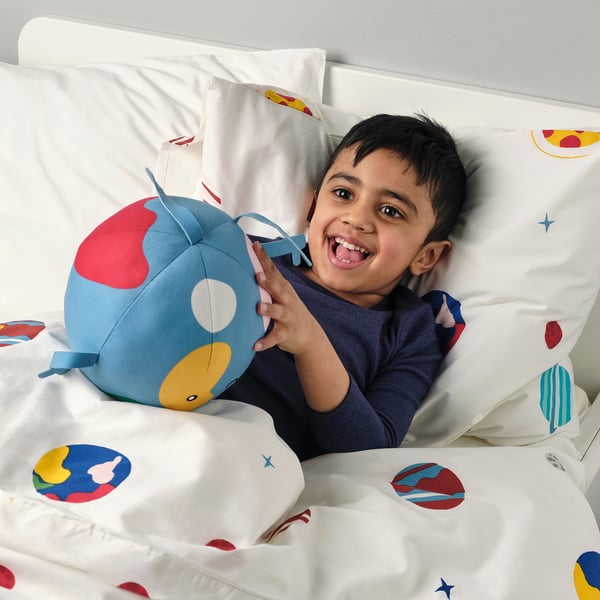 A child lying on a space-themed bed, hugging and playing with a colorful, soft, spherical toy.