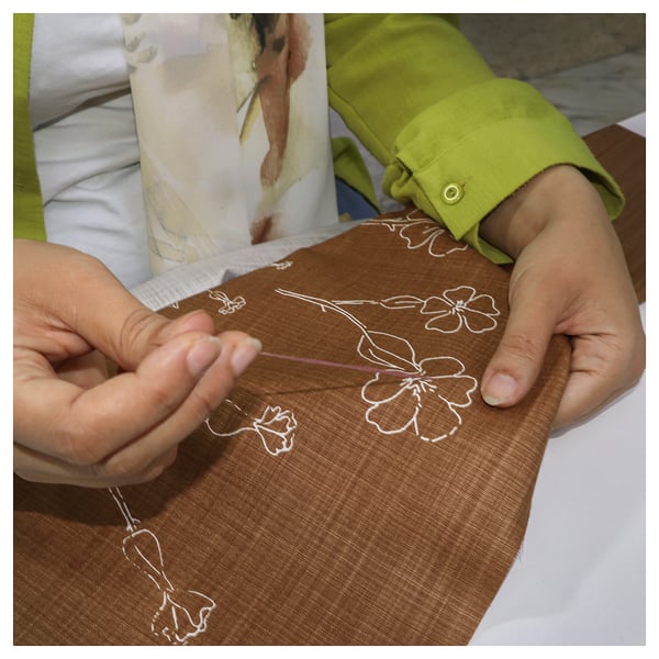 In hands are seen embroidering a brown cushion cover with white floral patterns. The fabric is cotton, and the design delicate hand-stitched details.