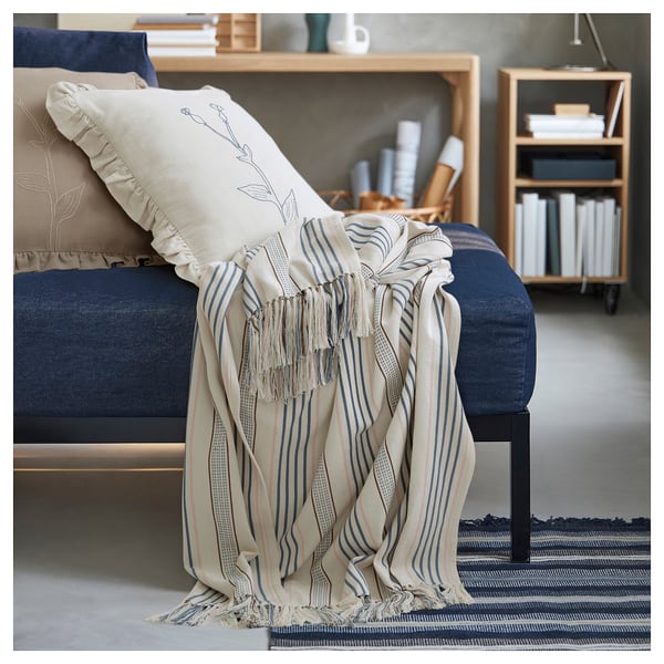 A cozy living room with a blue sofa, decorated with a cream and blue striped throw blanket and pillows. A wooden bookshelf is in the background.