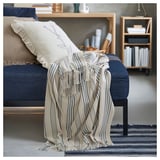 A cozy living room with a blue sofa, decorated with a cream and blue striped throw blanket and pillows. A wooden bookshelf is in the background.