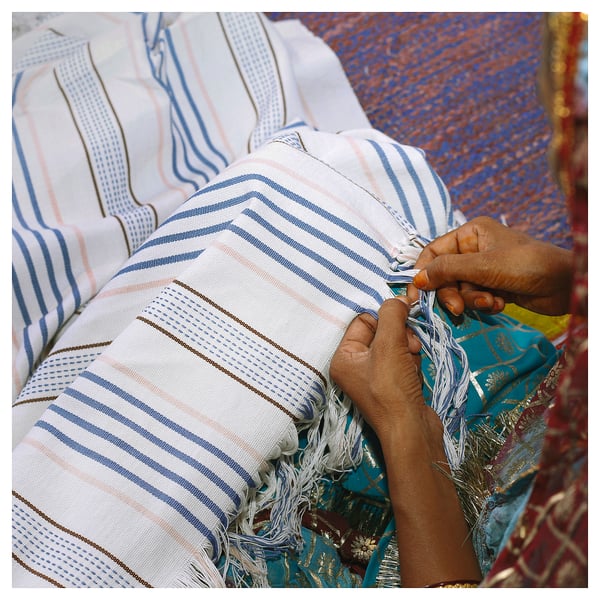 A person weaves a striped white and blue cotton throw with fringes, showcasing ÅKERNEJLIKA design.