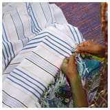 A person weaves a striped white and blue cotton throw with fringes, showcasing ÅKERNEJLIKA design.