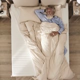 Sleeping bearded man in blue pajamas on a beige bed with pillows and nightstands.