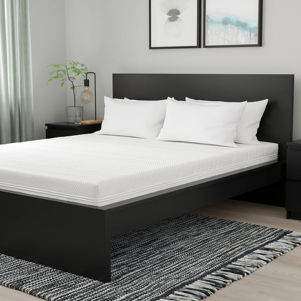 Modern black bed with firm ÅNNELAND white mattress, pillows, nightstand with lamp & plant. Rug, wall art, light grey curtain.