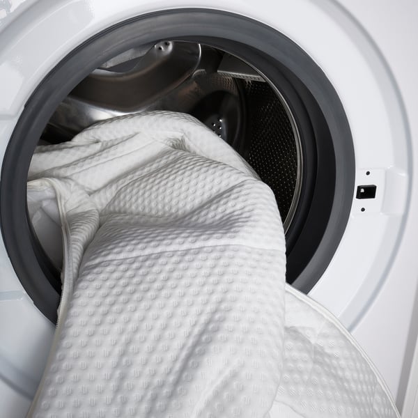 Image shows ÅNNELAND hybrid mattress being placed in a washing machine, highlighting its machine-washable cover for easy cleaning.