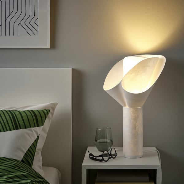 Bedside table with BÄCKNATE lamp, modern white design. Lamp stands tall near green pillows, glasses, and a wine glass. Soft, flower-like shade.