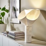 Country living room with white BÄCKNATE lamp and flower shade on sideboard, surrounded by books and a plant for a cozy feel.