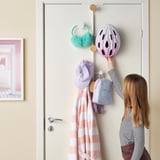 Image shows a child adjusting a pink helmet on a door-mounted coat hanger. The hanger holds a striped blanket, a hat, a backpack, and earmuffs.
