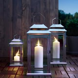 Three lit lanterns with candles, two with glowing flames, placed outdoors on a wooden surface.