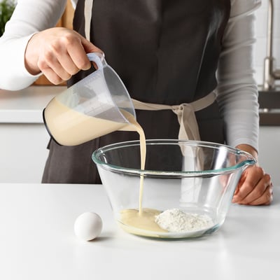 Person pours liquid from a measuring cup into a mixing bowl. The cup has multiple measurement scales visible.