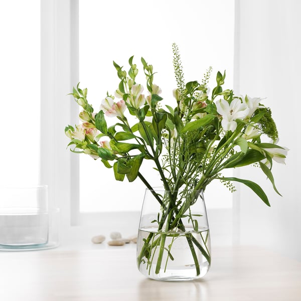 A clear glass vase with white flowers and green leaves sitting on a white table, holding water for the flowers.