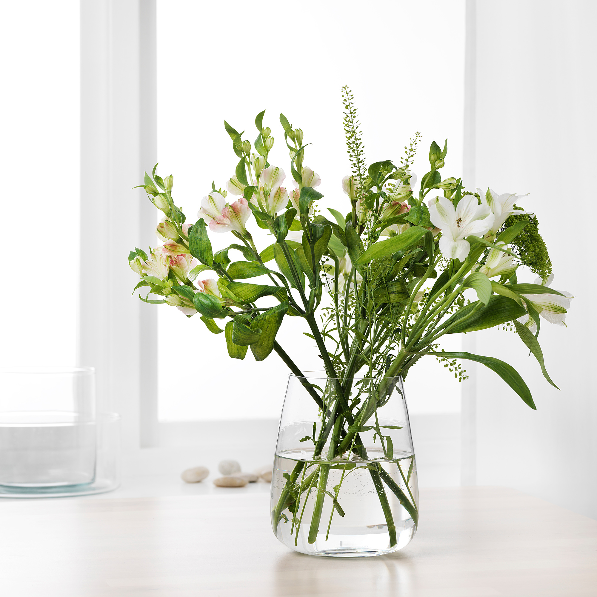 A clear glass vase with white flowers and green leaves sitting on a white table, holding water for the flowers.