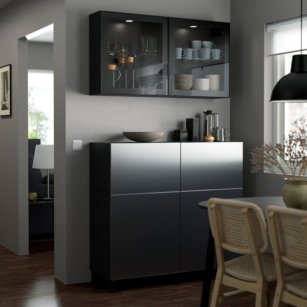 Modern dining area with gray BESTÅ storage. Glass cabinet displays dishes. Wicker chair, pendant light, and wooden floor. Combat-zone wall color.