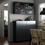 Modern dining area with gray BESTÅ storage. Glass cabinet displays dishes. Wicker chair, pendant light, and wooden floor. Combat-zone wall color.
