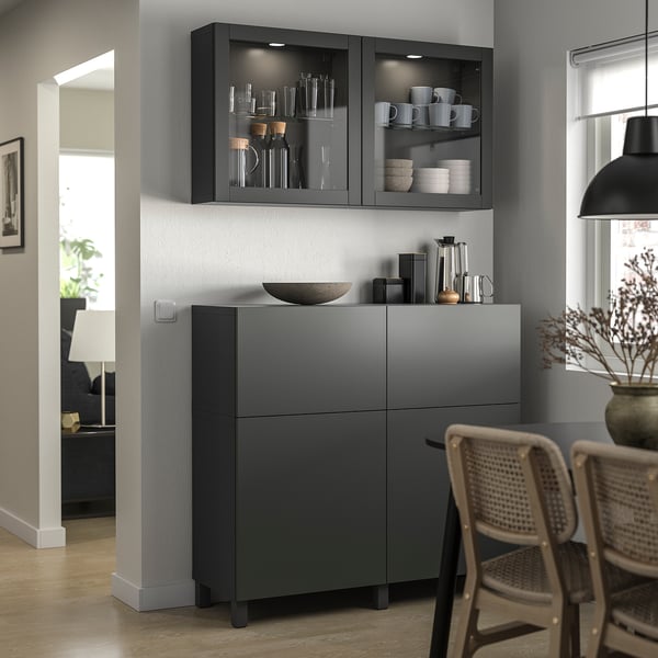 Modern dining area with dark gray BESTÅ storage units. Shelves hold glassware and dishes, showcased behind glass doors.