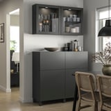 Modern dining area with dark gray BESTÅ storage units. Shelves hold glassware and dishes, showcased behind glass doors.