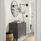 Modern entryway with black BESTÅ storage unit, round mirror, wall lamps, large vase with branches, books, and woven basket.