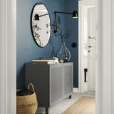 Modern hallway with gray BESTÅ storage unit, featuring woven basket, blue vase, books, and circular mirror on blue wall.
