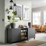 Black BESTÅ storage unit with shelves, holding plant, books, and bowl. Modern, minimalist design in a grey living room with orange couch and artwork.