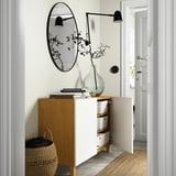 Modern entryway with white and wood cabinet. Open door shows storage boxes. Mirror, wall light, vase with branches, and woven basket are also present.