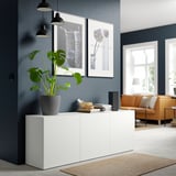 Modern living room with BESTÅ sideboard, books, plant, and art. Orange couch nearby.