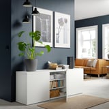 Modern living room with white BESTÅ unit, decorated with books, photos, lamps, and a large plant, orange sofa, navy wall.
