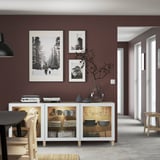 White BESTÅ sideboard with glass doors, displaying dishes and vases, under three framed artworks on a mocha wall.