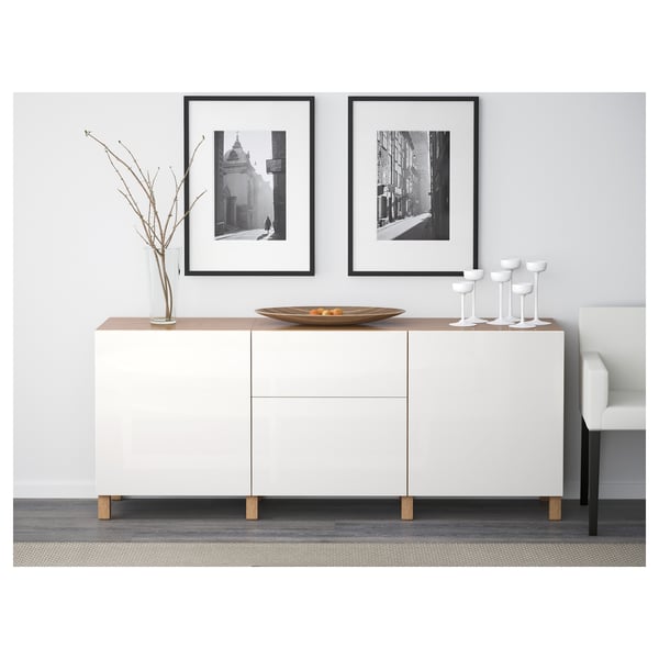 White sideboard with wooden legs, displaying vase, bowl, and glasses. Framed black-and-white photos hang above. Modern, minimalist living room setup.
