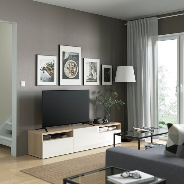 Modern living room with BESTÅ tv bench, black tv, white drawers, gray curtains. Artwork above tv, plants, and lamp beside. Sofa in front.
