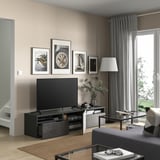 Modern living room with beige walls, black BESTÅ tv bench, framed art, glass coffee table, grey sofa, large window with grey curtains.