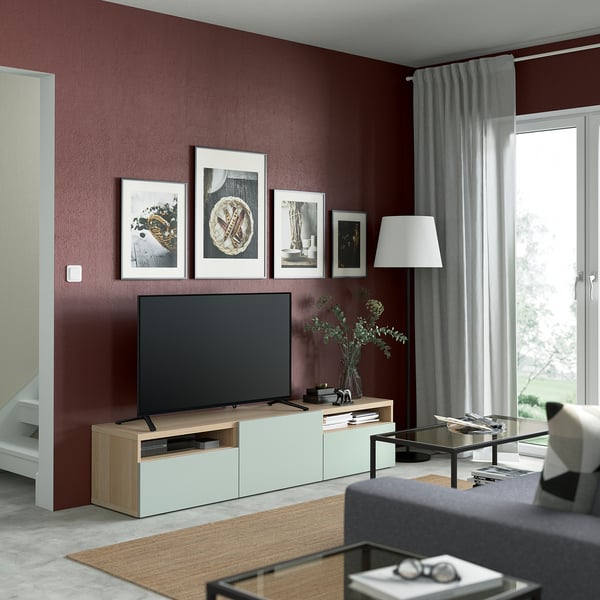 Modern living room with tv, dark red wall, framed pictures, lamp, grey curtains, coffee table items, grey sofa.
