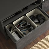 Open gray BESTÅ tv drawer, white dividers storing black gaming controllers, headphones, remotes.