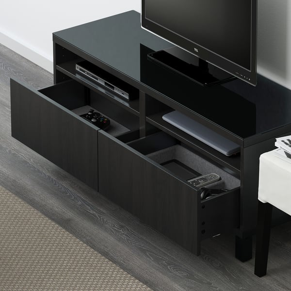Black tv stand with drawers, holding controllers and devices, with glass top and cable management at the back.