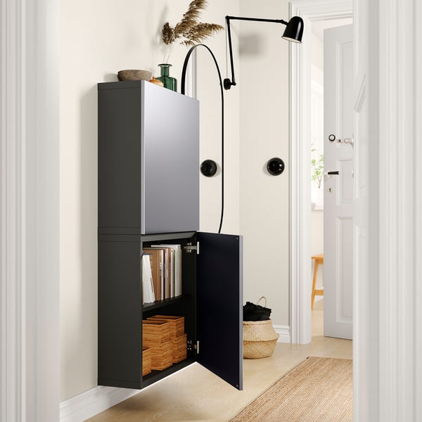 Modern hall: black BESTÅ wall cabinet, open showing books & boxes, next to black wall lamp on white walls.