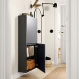 Modern hall: black BESTÅ wall cabinet, open showing books & boxes, next to black wall lamp on white walls.