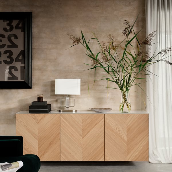 Modern living room with light wood BESTÅ sideboard, white lamp, tall plant in clear glass vase, green chair, black picture frame, and beige curtains.