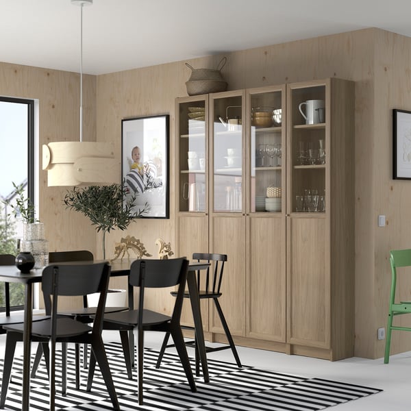 Modern dining area with large wooden BILLY bookcase, black chairs, striped rug. Light wood, glass doors showcase dishes. Simple, clean design.